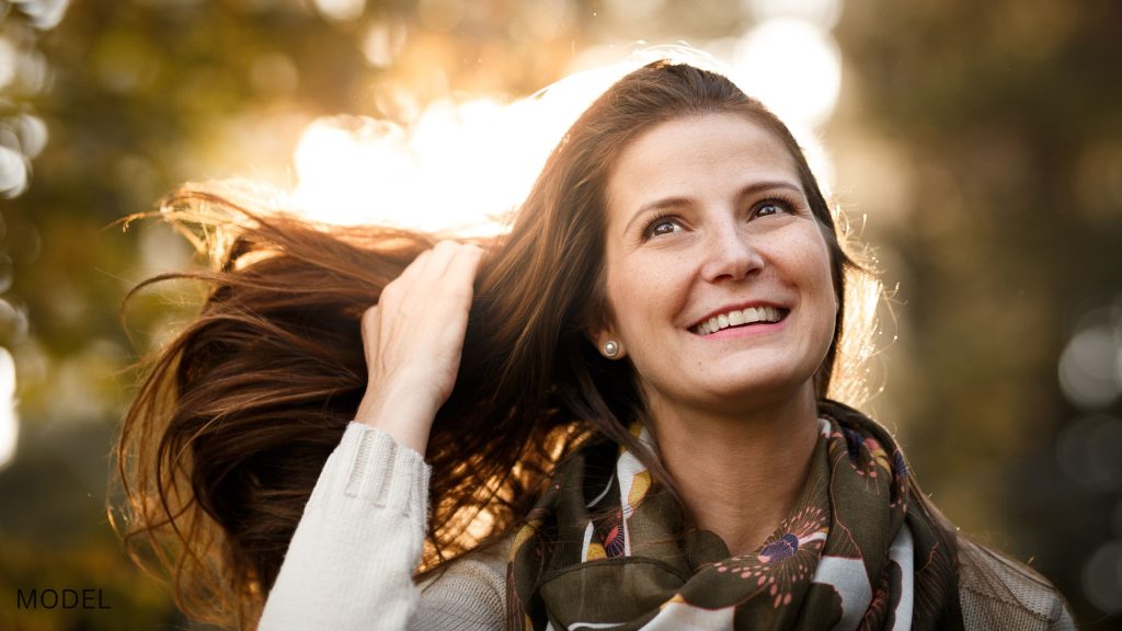 A woman with beautiful hair stands outside while holding her hair has it blows in the wind. (Model)